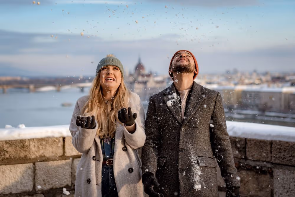 Smiling young woman and man wearing winter coats and hats playing with falling snow outdoors near a river.