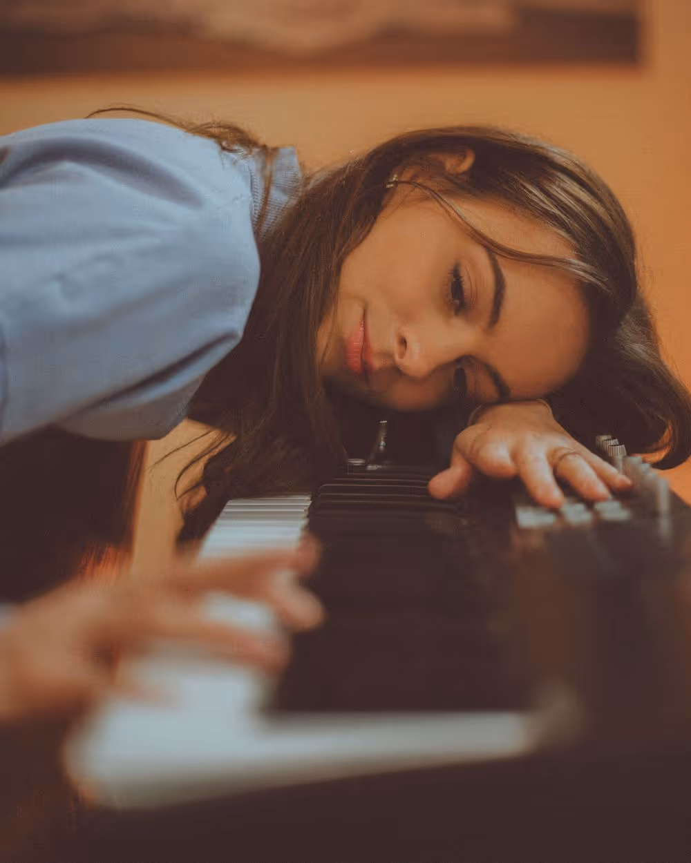 Woman resting her head and hand on the keys of a piano keyboard, with a focused and calm expression.