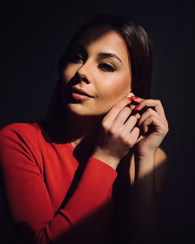 Woman in a red top adjusting an earring with soft lighting creating shadows on her face.