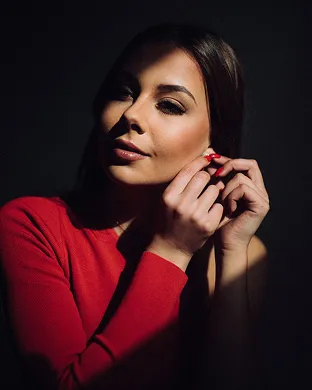 Woman in a red top adjusting an earring with soft light highlighting her face against a dark background.