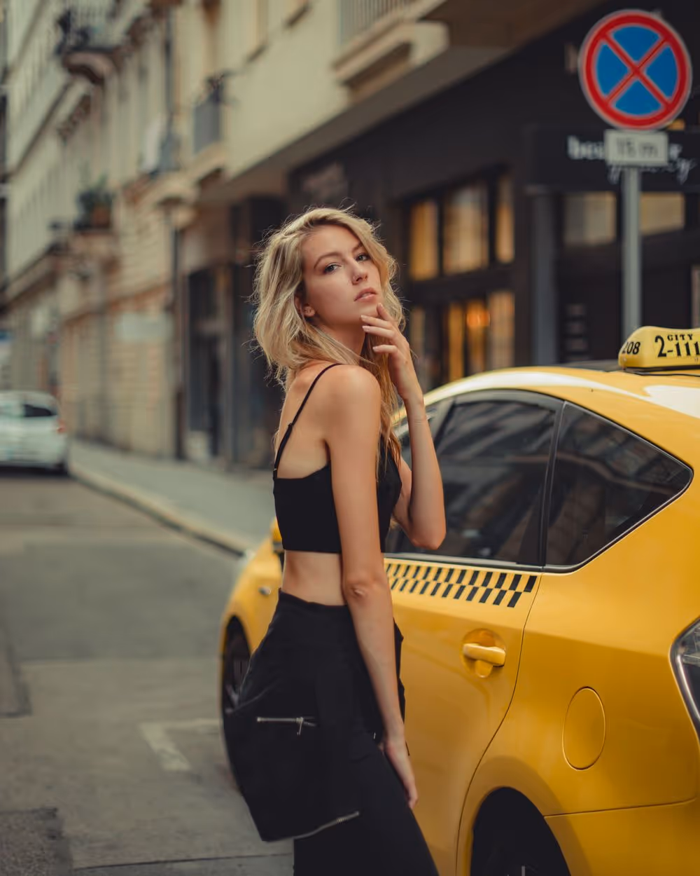 Blonde woman in black crop top and pants posing next to a yellow taxi on a city street.