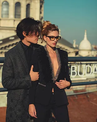 Fashionable man and woman standing outdoors near a railing with historic buildings in the background.