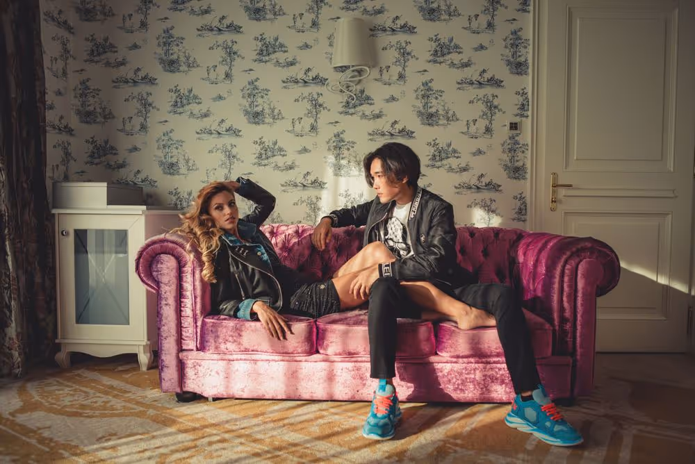 Man and woman sitting on a plush pink sofa in a room with patterned wallpaper, the woman reclining and the man looking at her.