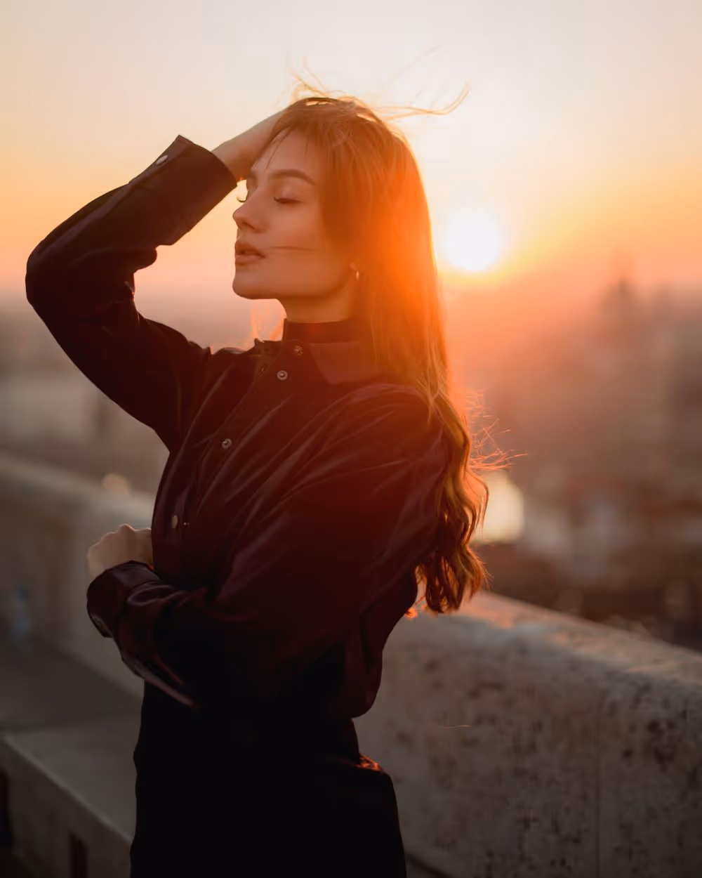 Woman in a black blouse with eyes closed, touching her hair at sunset on a rooftop.