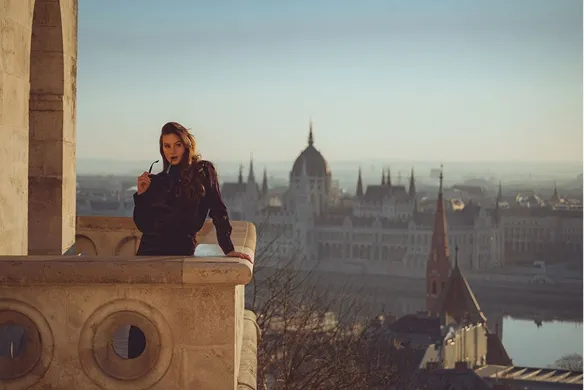Woman in dark clothing leaning on a stone balcony overlooking a cityscape with historic buildings and a river at sunset.