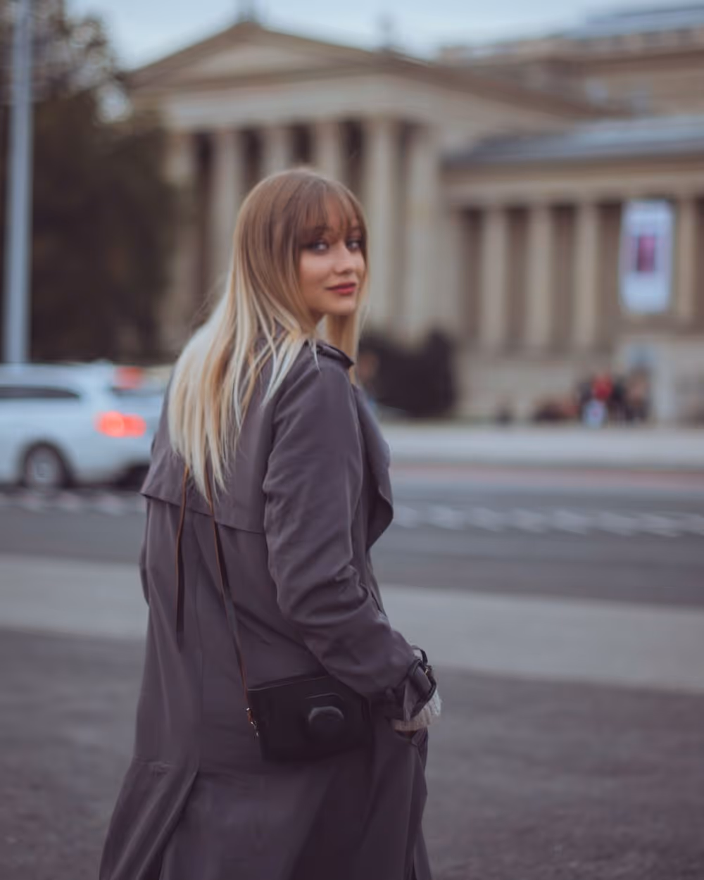 Young woman with long blonde hair wearing a dark coat and carrying a black purse, looking back while standing on a city street.