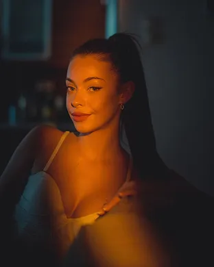 Young woman with long dark hair tied back, softly lit with warm light in a dim room.