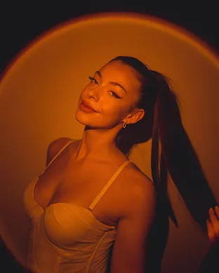 Young woman with long dark hair in a ponytail wearing a light-colored corset top, posing with a confident smile against a warm orange circular background.