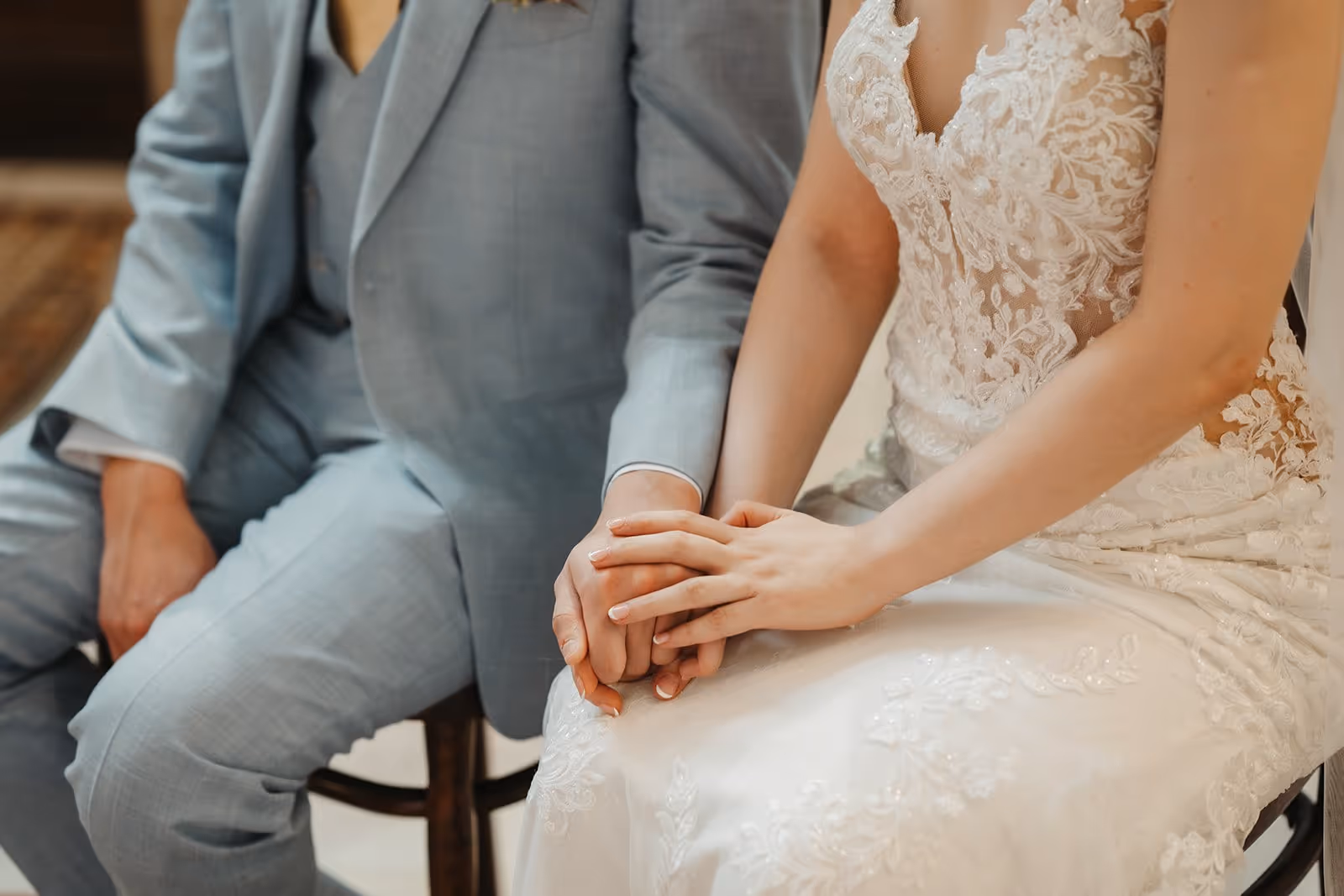 Bride in white lace dress and groom in light gray suit sitting close and holding hands.