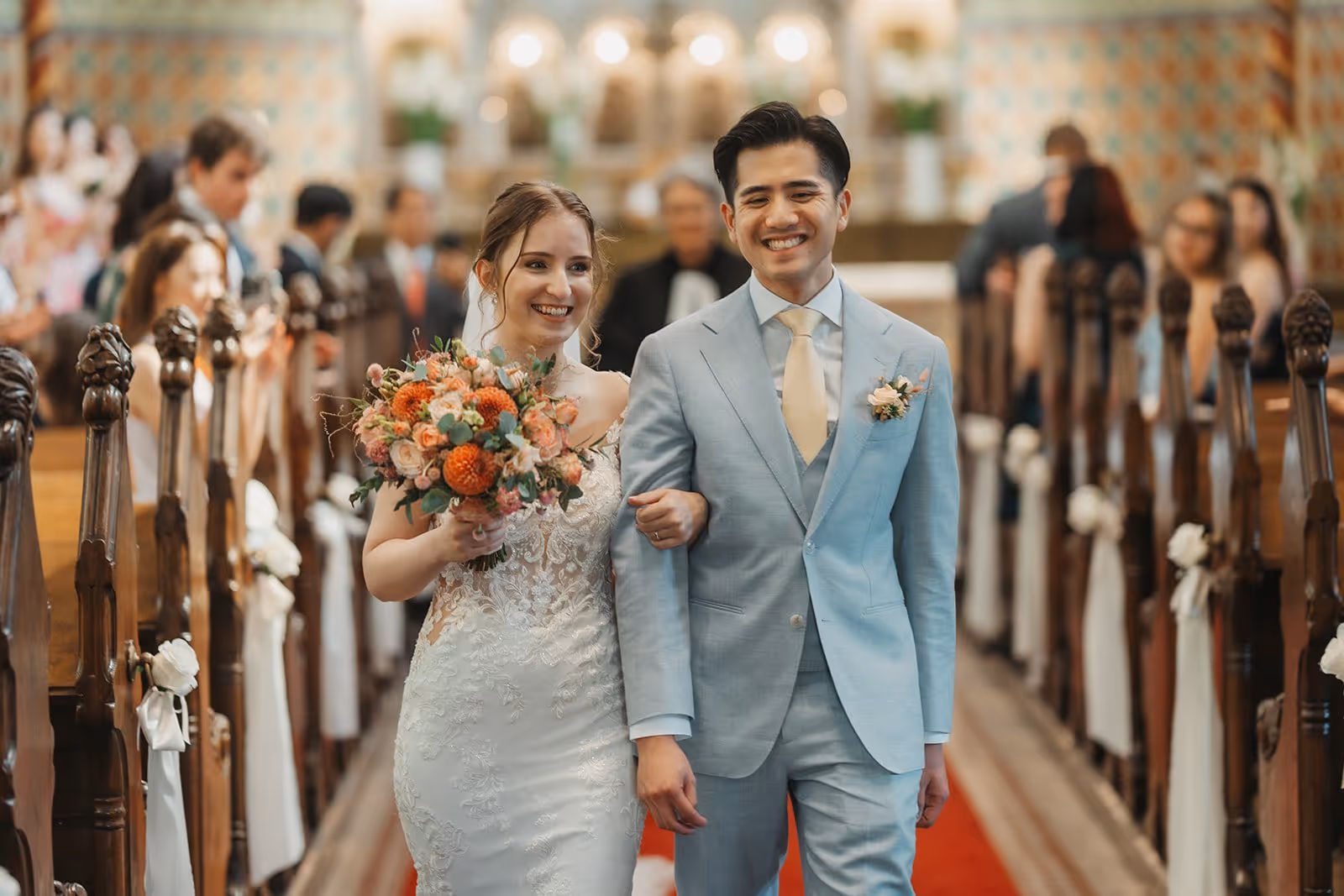 Bride in white lace dress holding a bouquet and groom in light blue suit walking arm in arm down a church aisle.