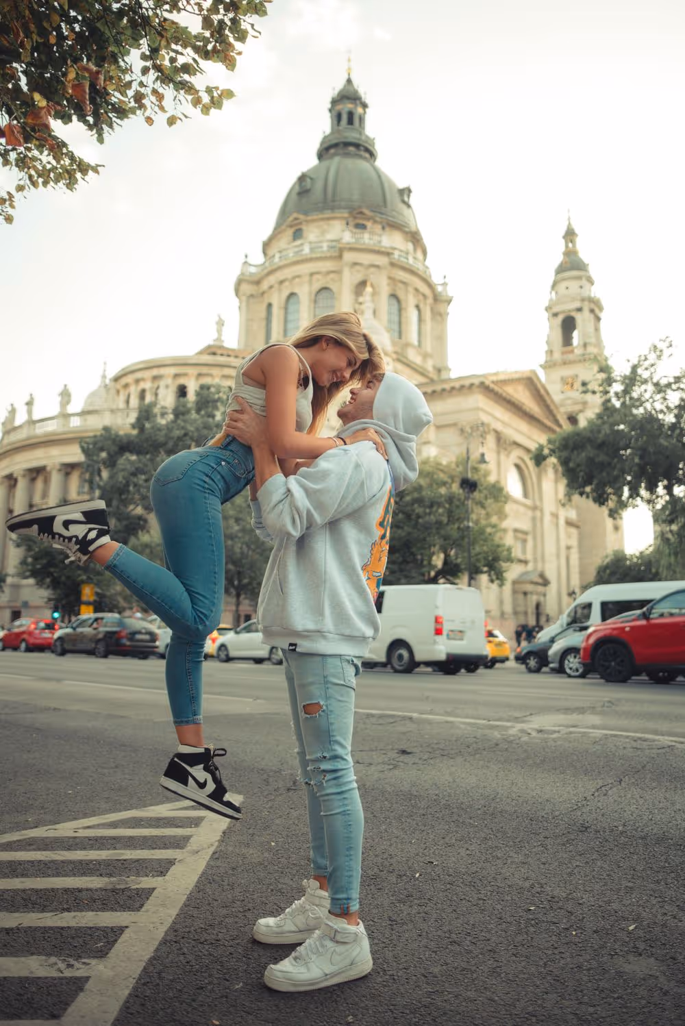 Young man in a hoodie lifting a smiling woman in jeans in front of a large historic building with a dome.