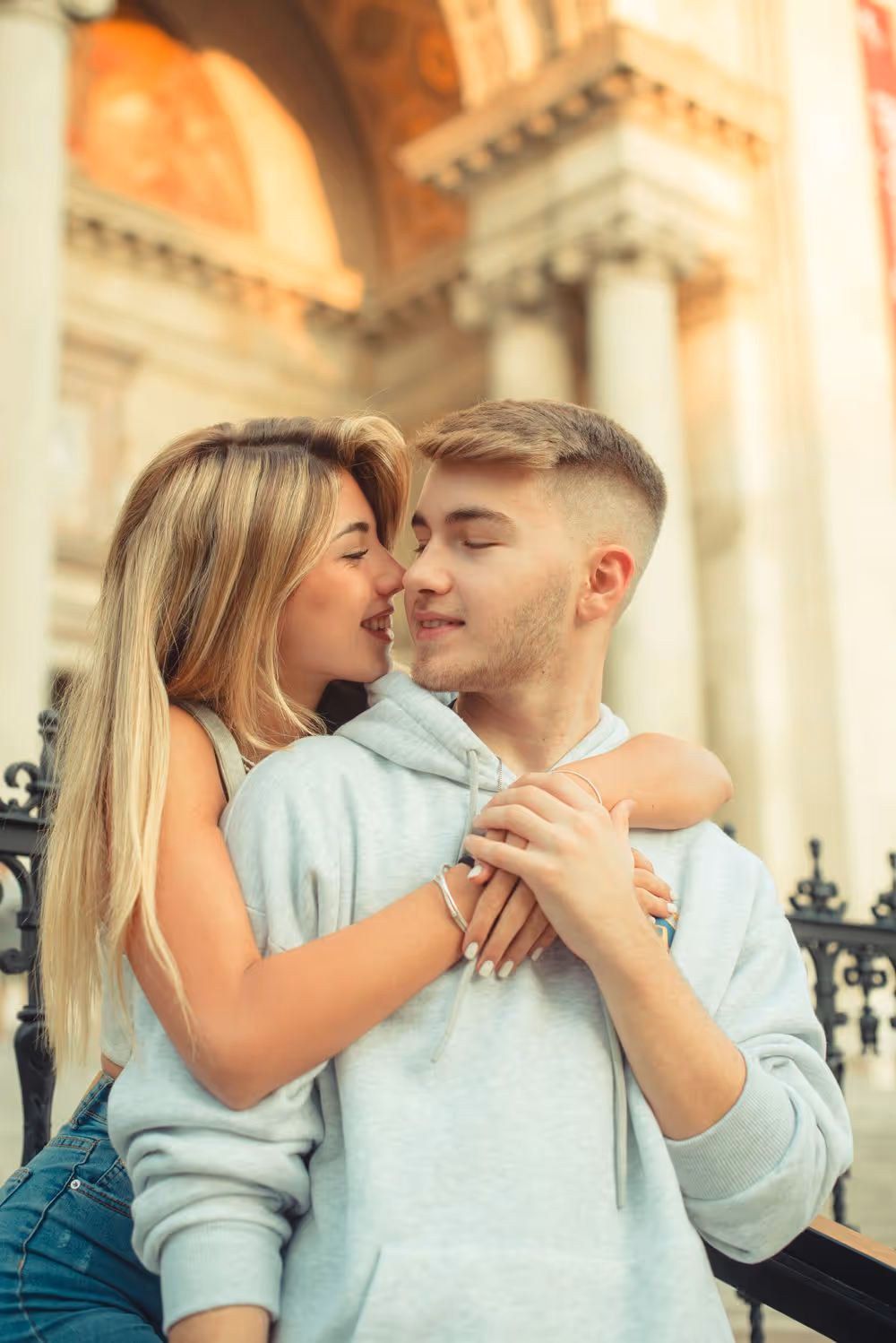 Young woman hugging a young man from behind while they smile affectionately at each other outdoors.