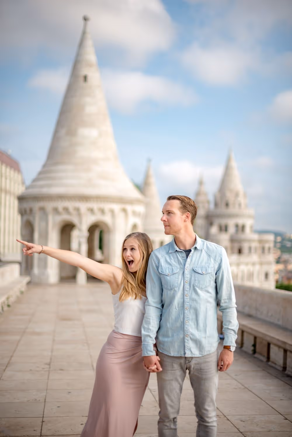 Couple holding hands with the woman excitedly pointing to the left, standing on a stone terrace with historic towers in the background.