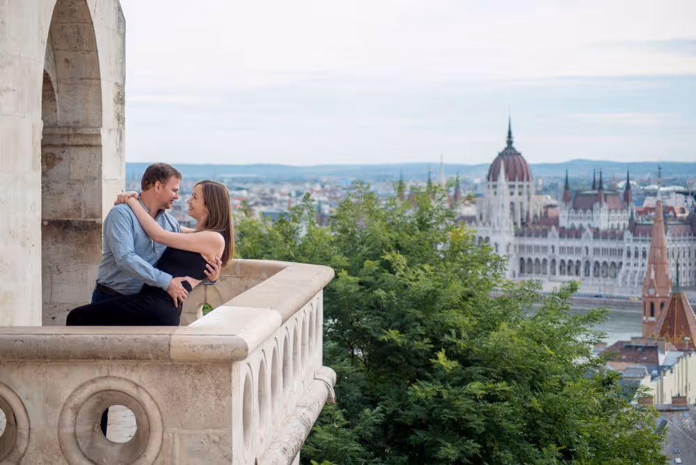 Couple embracing on a stone balcony overlooking a cityscape with historic buildings and greenery.