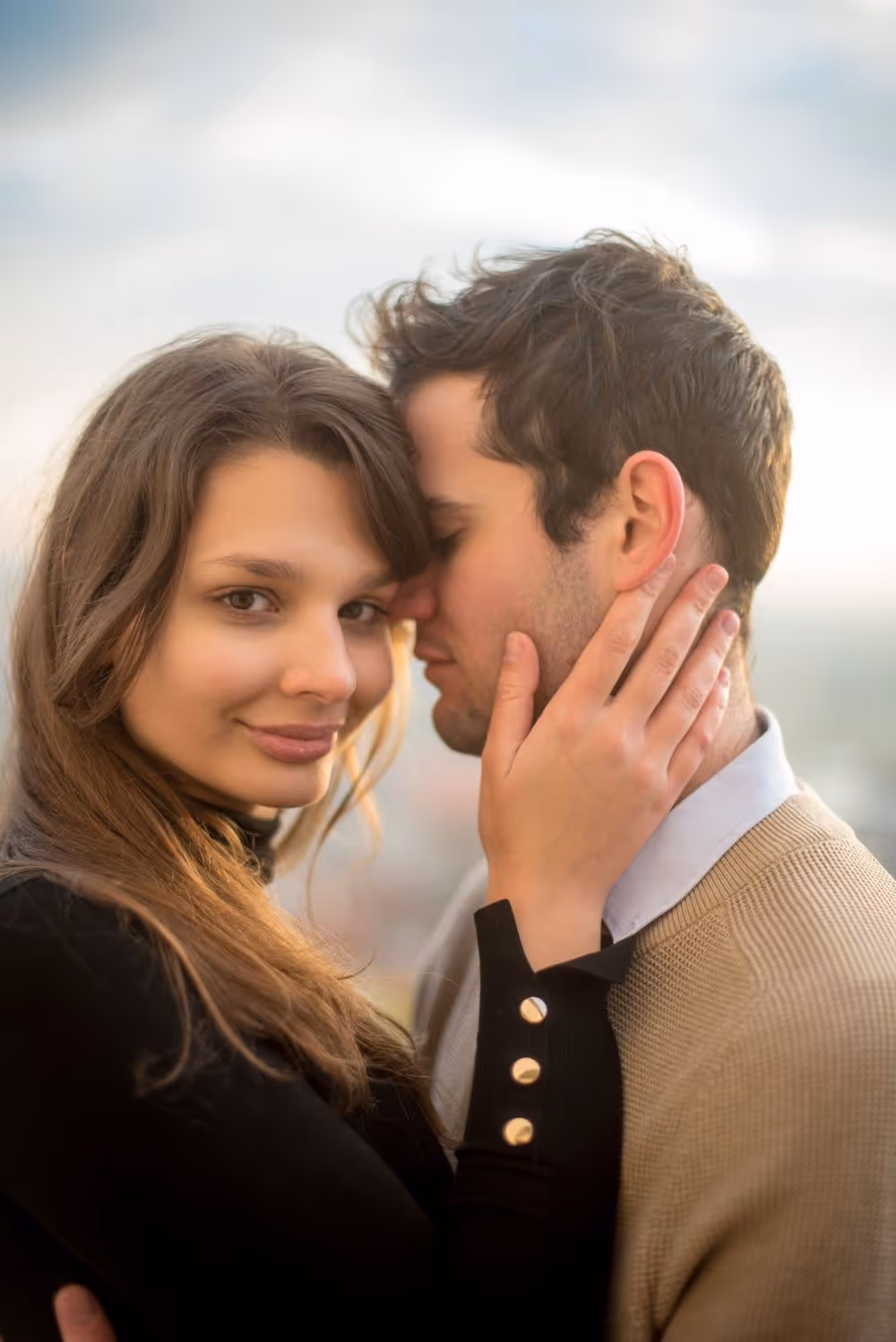 Couple embracing outdoors with woman gently holding man's face, looking at camera with a soft smile.