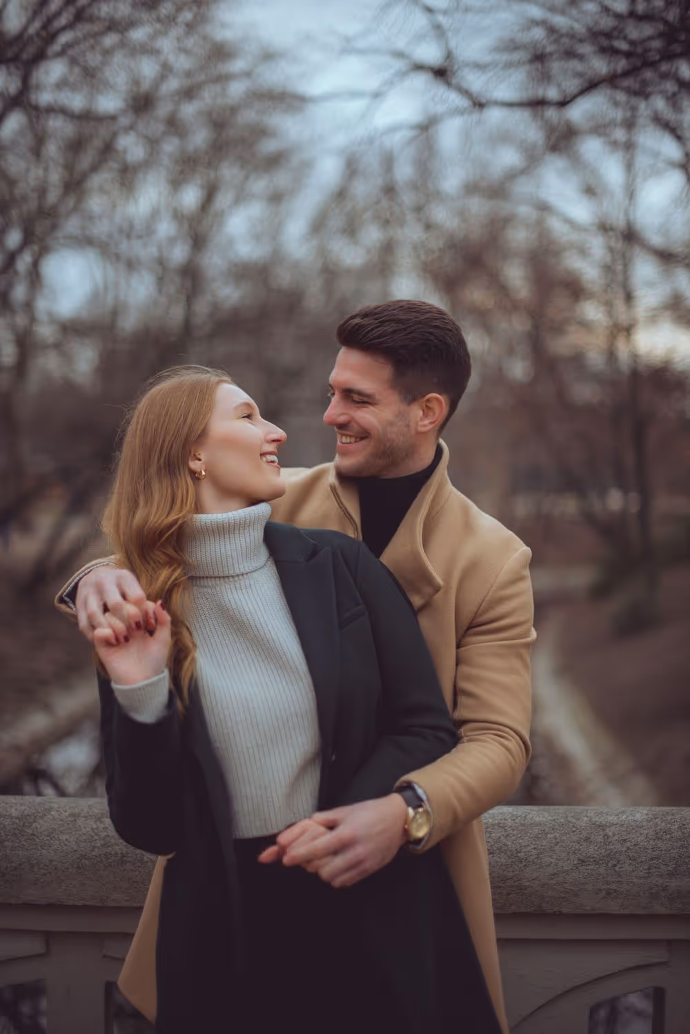 Smiling couple embracing on a stone bridge with bare trees in the background.