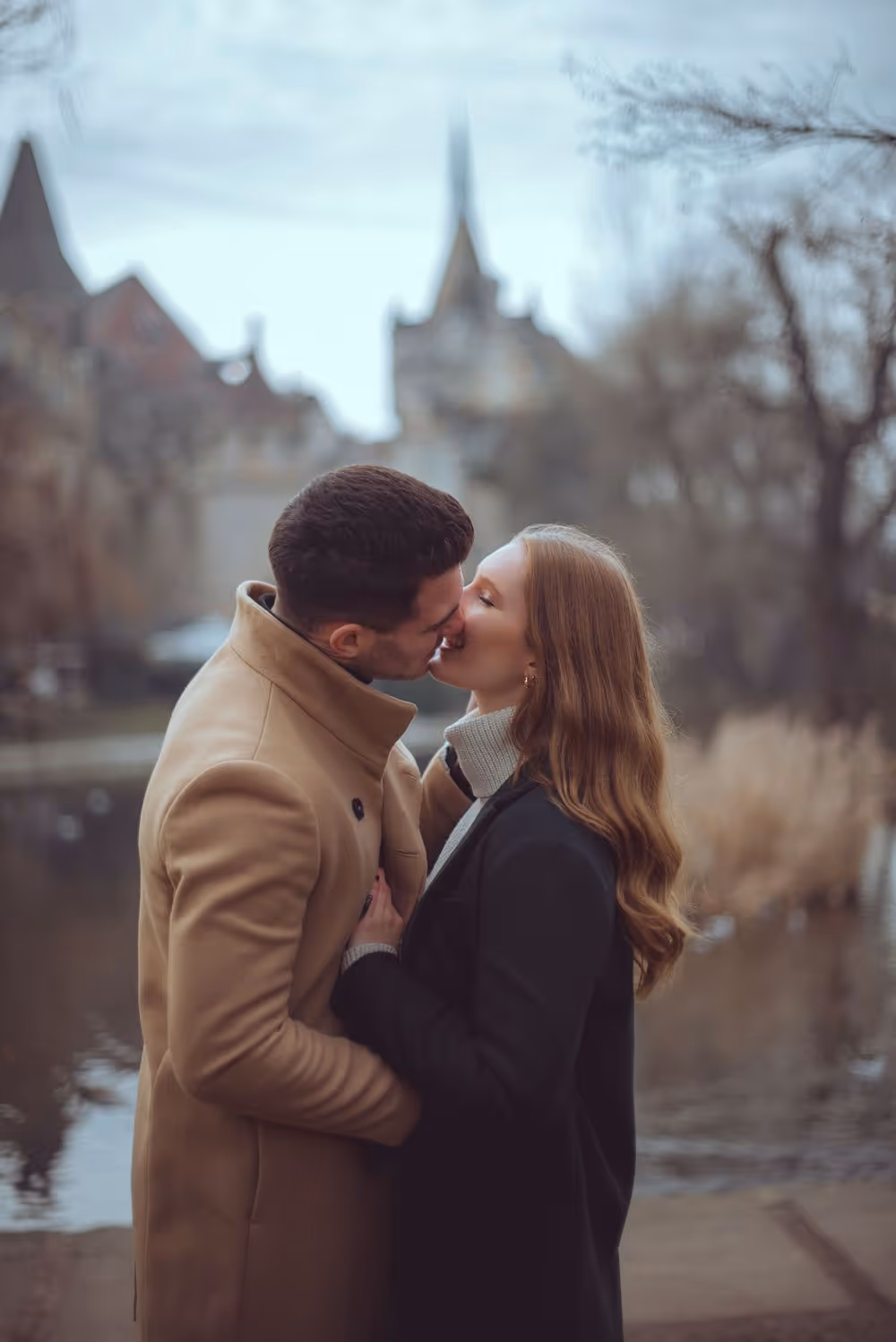 A couple kissing outdoors near a body of water with blurred buildings and trees in the background.