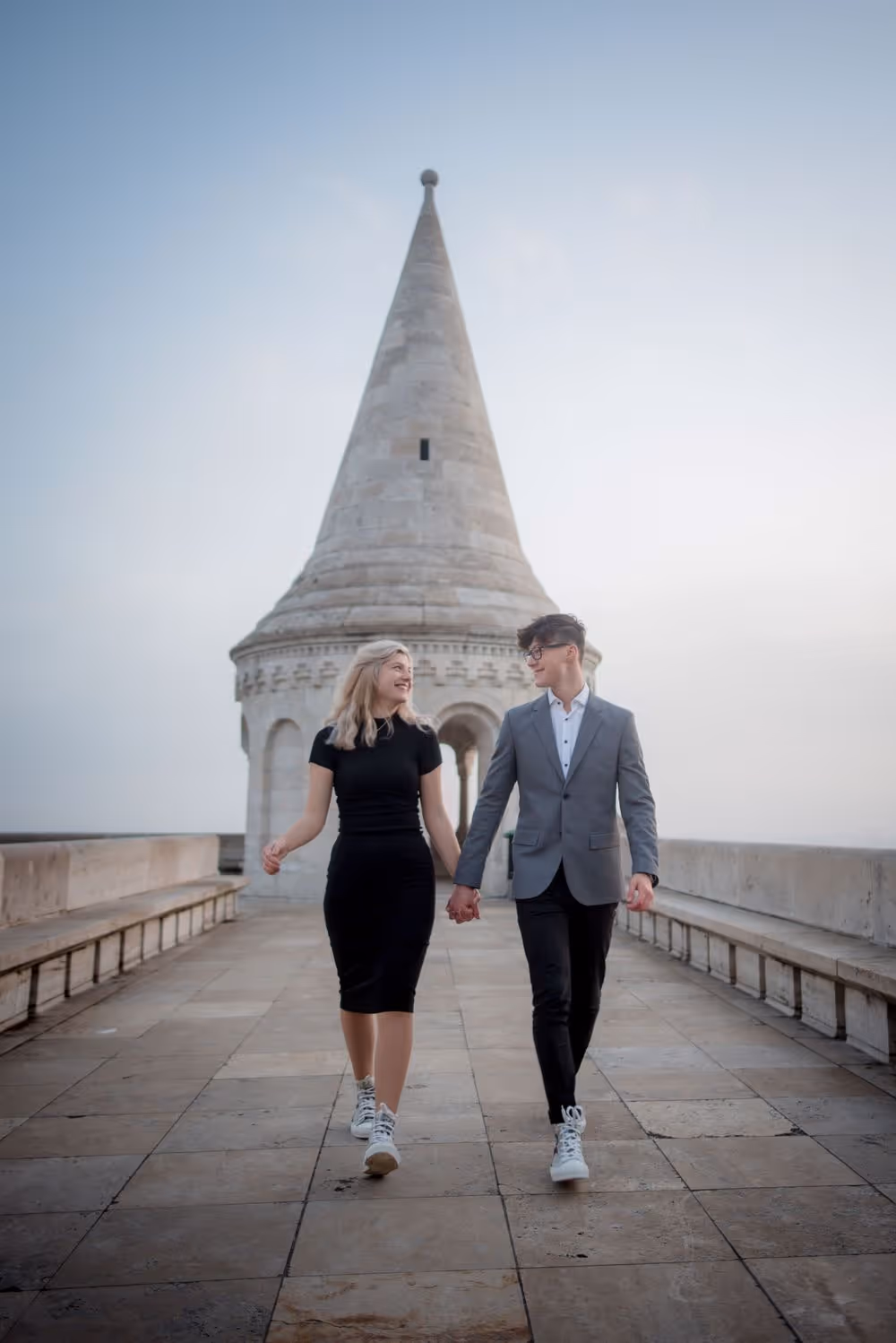 Couple holding hands and walking on a stone pathway with a tall, pointed stone tower in the background.