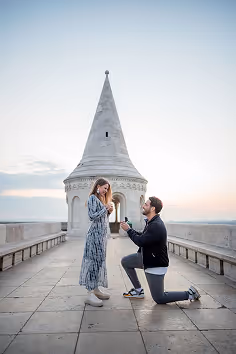 Man kneeling and proposing with a ring to a surprised woman in front of a historic stone tower at sunset.