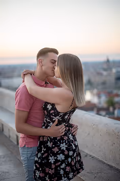 Young couple embracing and kissing on a balcony overlooking a city at sunset.