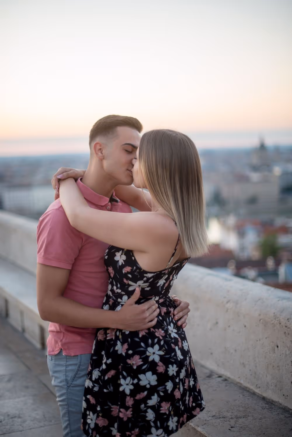 Young couple kissing outdoors on a terrace overlooking a city at sunset.