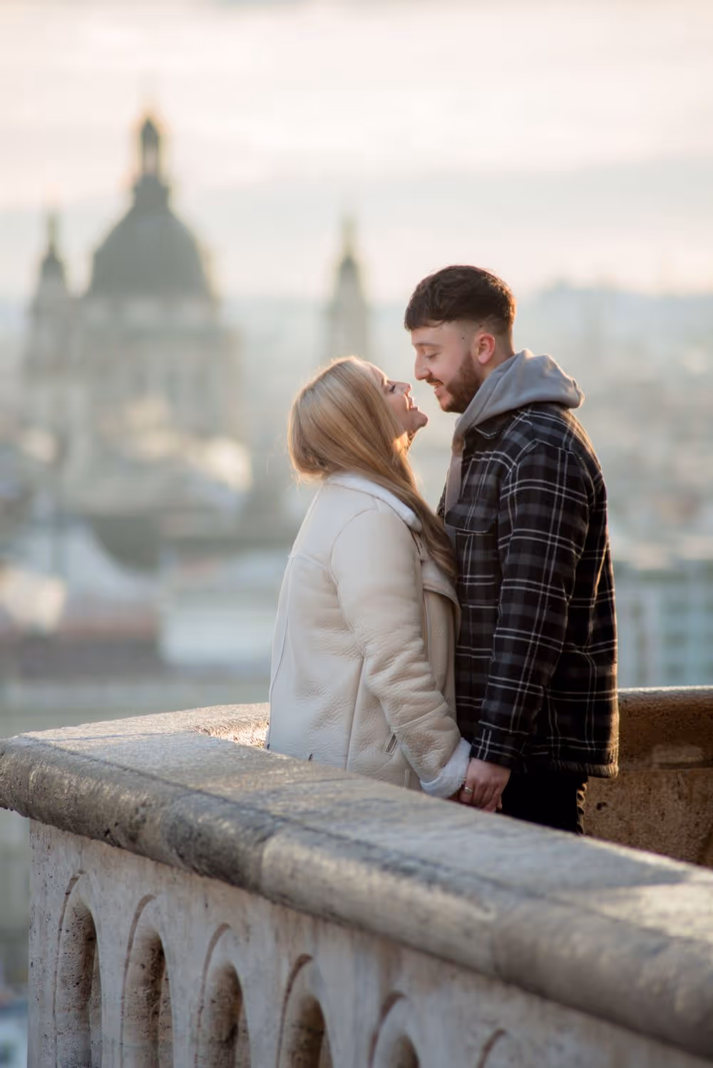 Couple holding hands and smiling at each other on a stone balcony overlooking a city with a blurred historic building in the background.