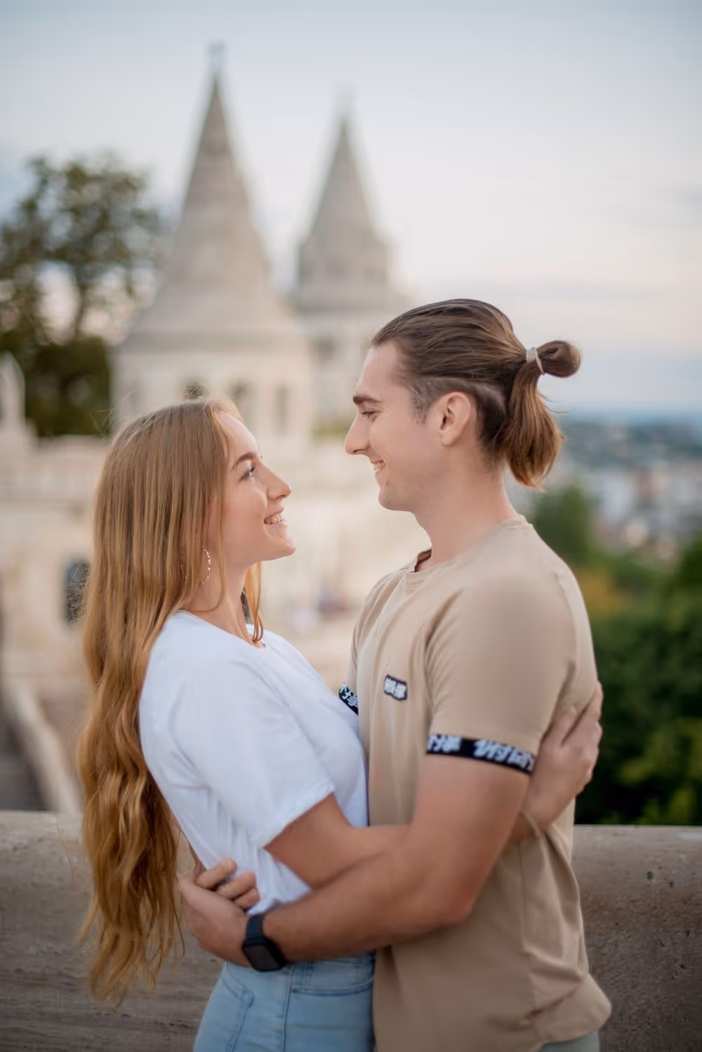Young couple smiling and embracing outdoors with blurred historic castle towers in background.