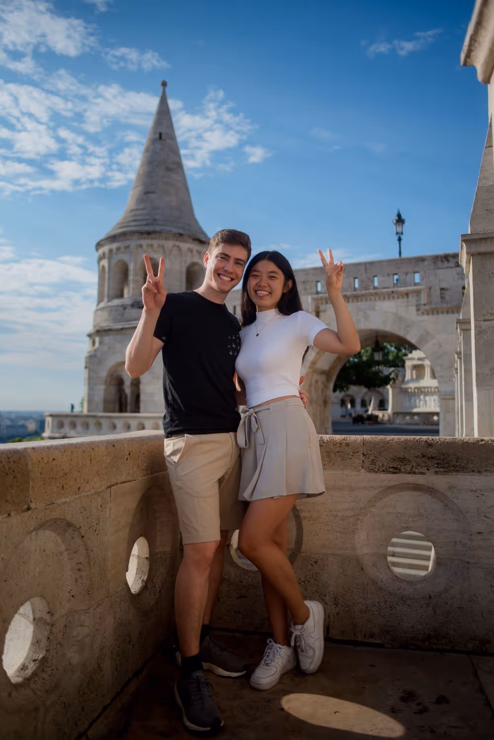Smiling young couple posing with peace signs on balcony of historic stone building under blue sky.