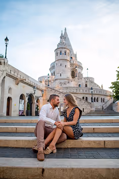Couple sitting on stone steps holding hands with Fisherman's Bastion tower in the background under a blue sky.
