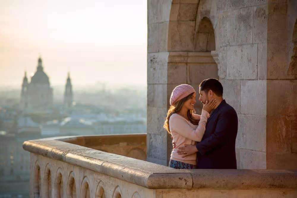 Couple embracing and smiling on a stone balcony overlooking a city with historic buildings at sunset.