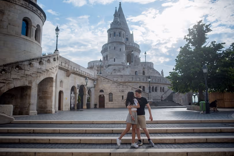 Couple embracing on stone steps in front of a historic castle-like building under a partly cloudy sky.