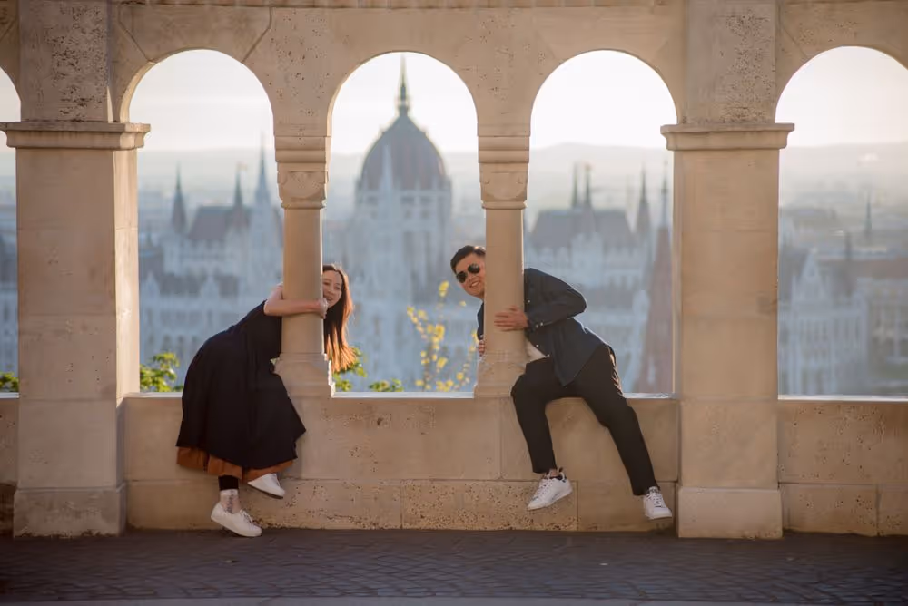 A man and a woman playfully leaning from stone arches with a blurred cityscape and a domed building in the background.