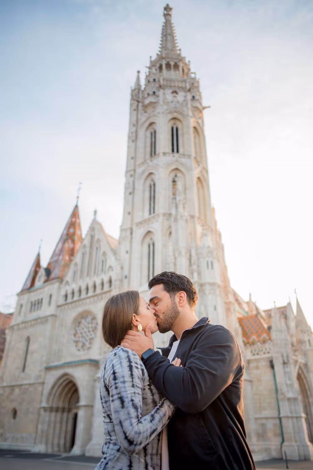 Couple kissing in front of a tall, ornate stone church tower with a clear sky.