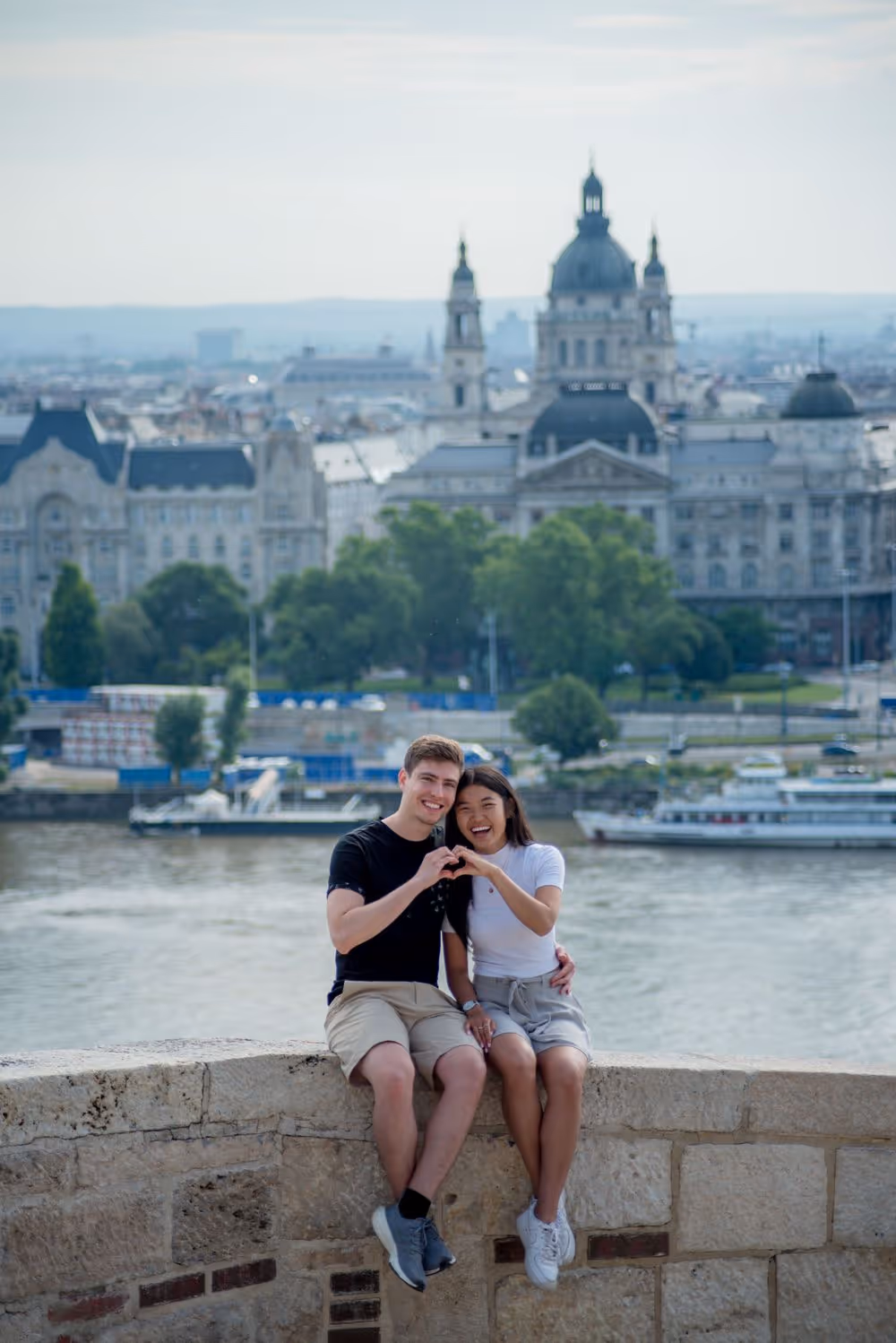 Smiling young couple sitting on a stone ledge near a river, forming a heart shape with their hands, with a cityscape and historic buildings in the background.