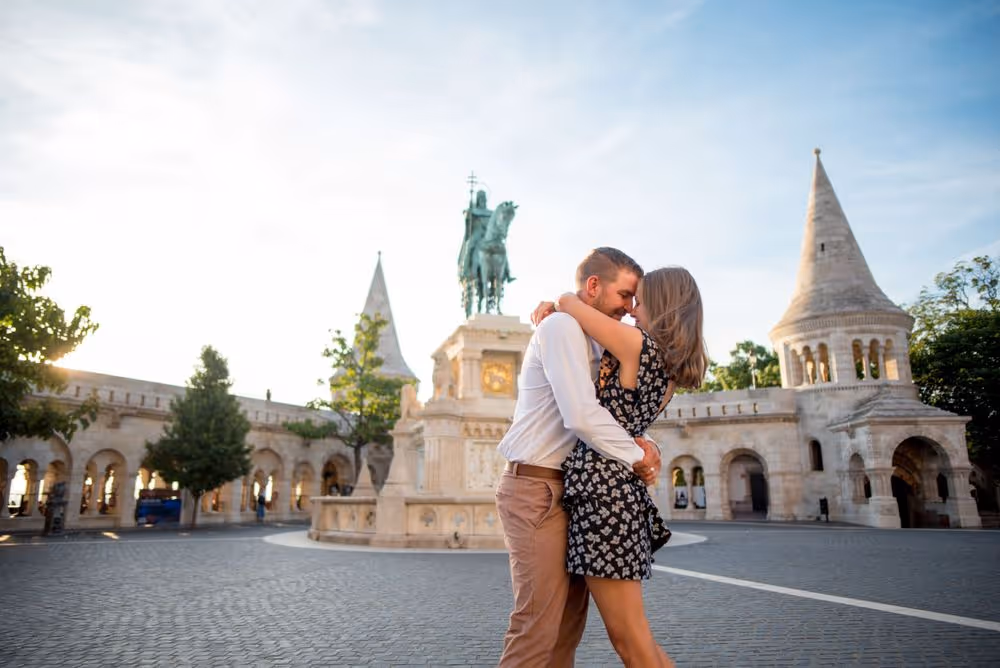 Couple embracing and smiling in front of a historic stone monument with arched buildings and towers during daytime.