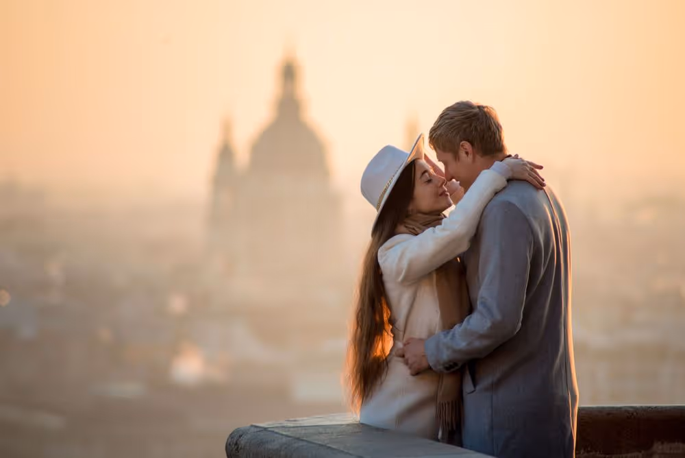 Couple embracing tenderly on a rooftop with a blurred cityscape and cathedral in the background at sunset.