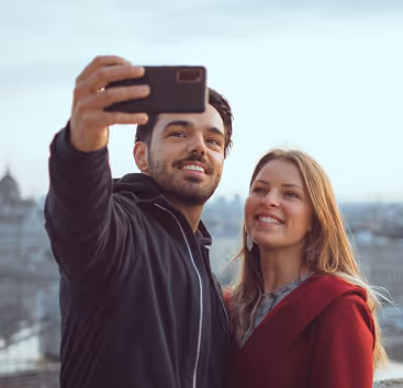 Smiling couple taking a selfie outdoors with a cityscape in the background.