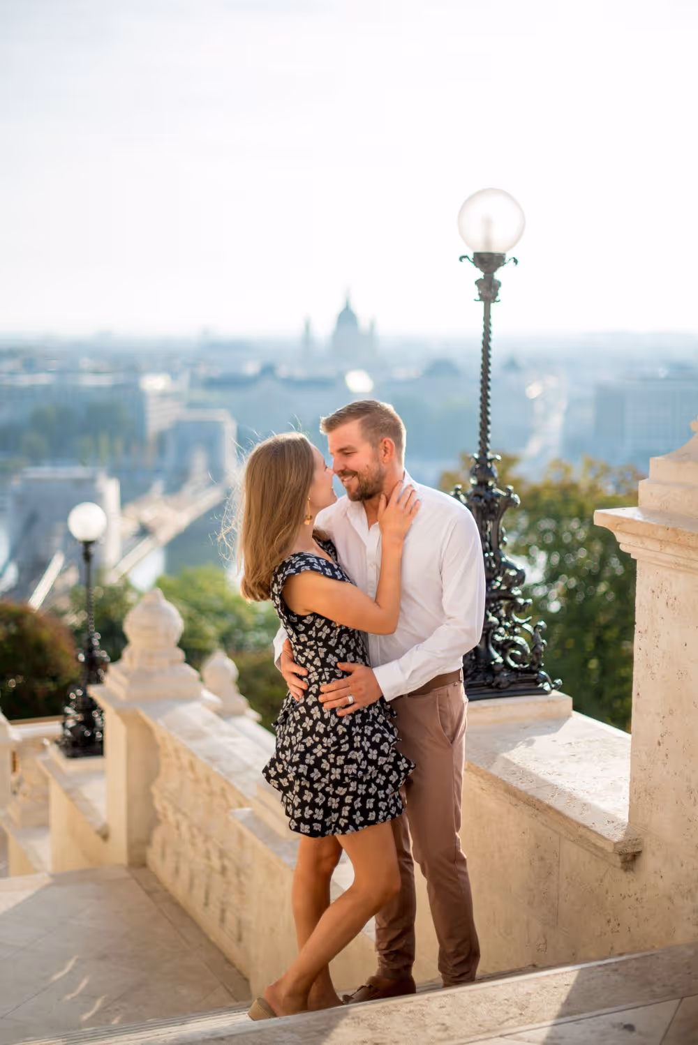 Couple embracing on outdoor stone staircase with cityscape in the background during daytime.