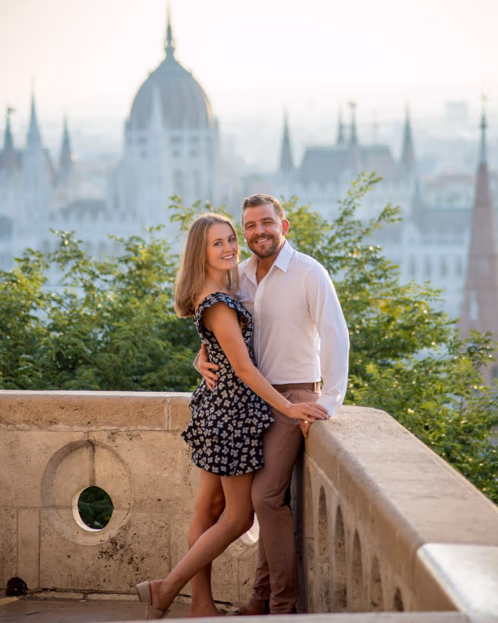 Smiling couple embracing on a stone terrace with greenery and blurred ornate building in the background.