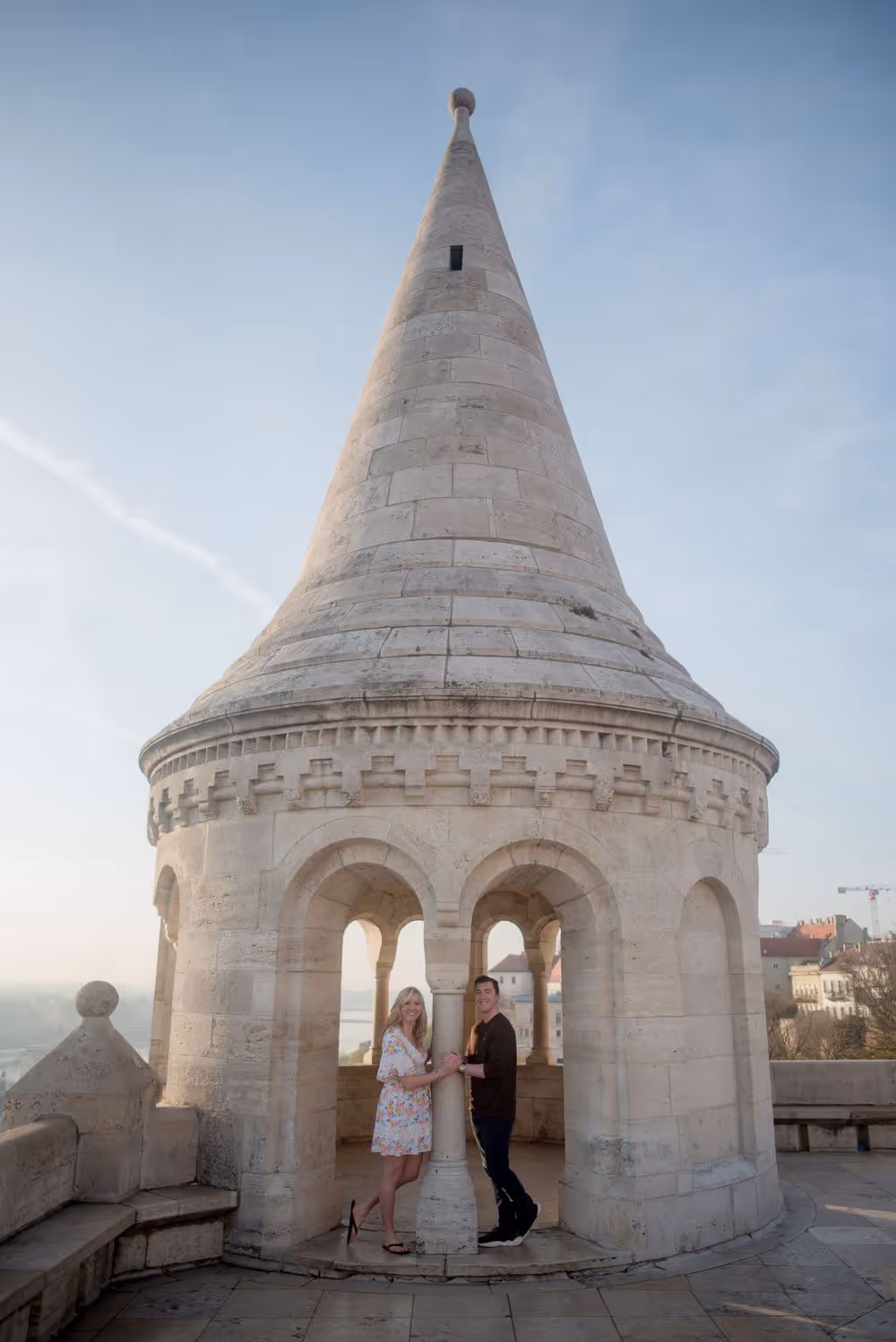 A couple holding hands and smiling inside a round stone tower with arched openings under a clear sky.