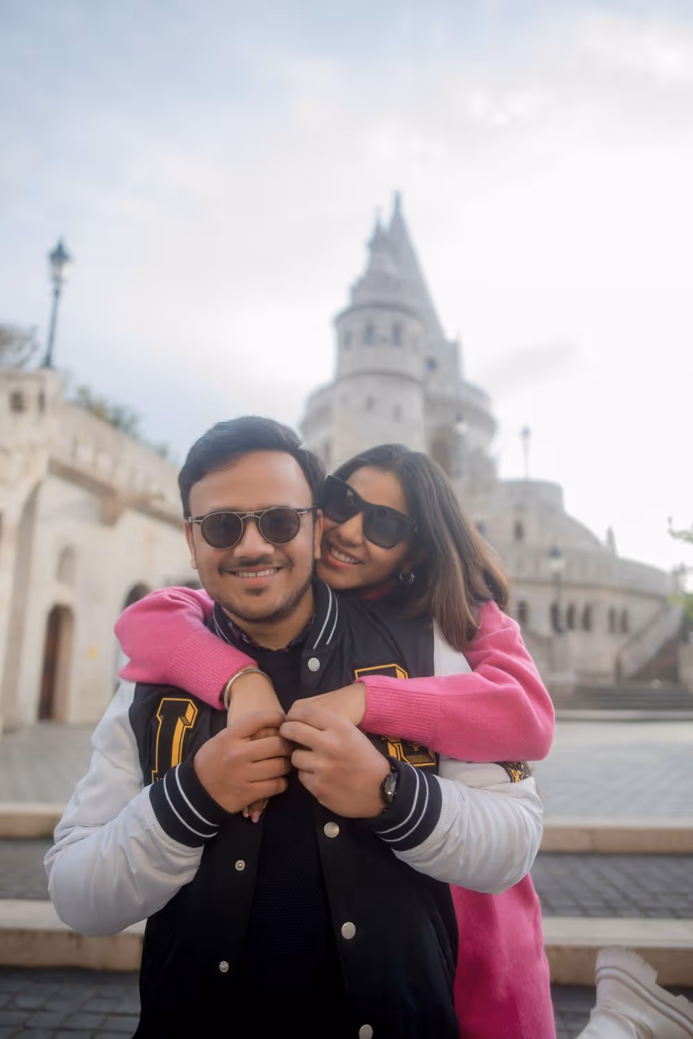 Smiling couple wearing sunglasses embraces in front of a historic stone tower with arches and stairs.