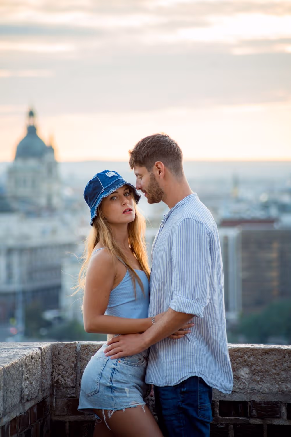 Young couple embracing on a rooftop with a cityscape and church dome in the background at sunset.