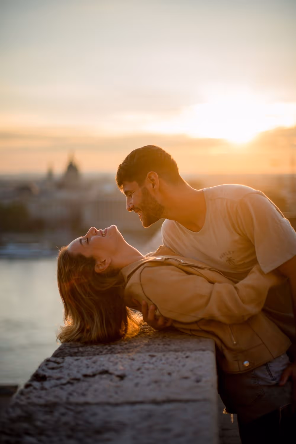 Couple sharing a joyful moment leaning over a stone ledge at sunset by a waterfront.