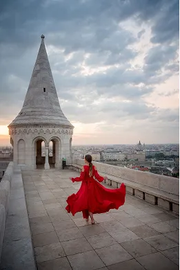 Woman in flowing red dress walking on stone terrace near a historic tower at sunset under a cloudy sky.