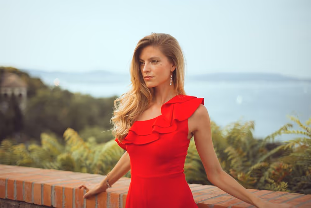 Woman in a red dress with ruffles standing outdoors by a brick wall with greenery and a body of water in the background.