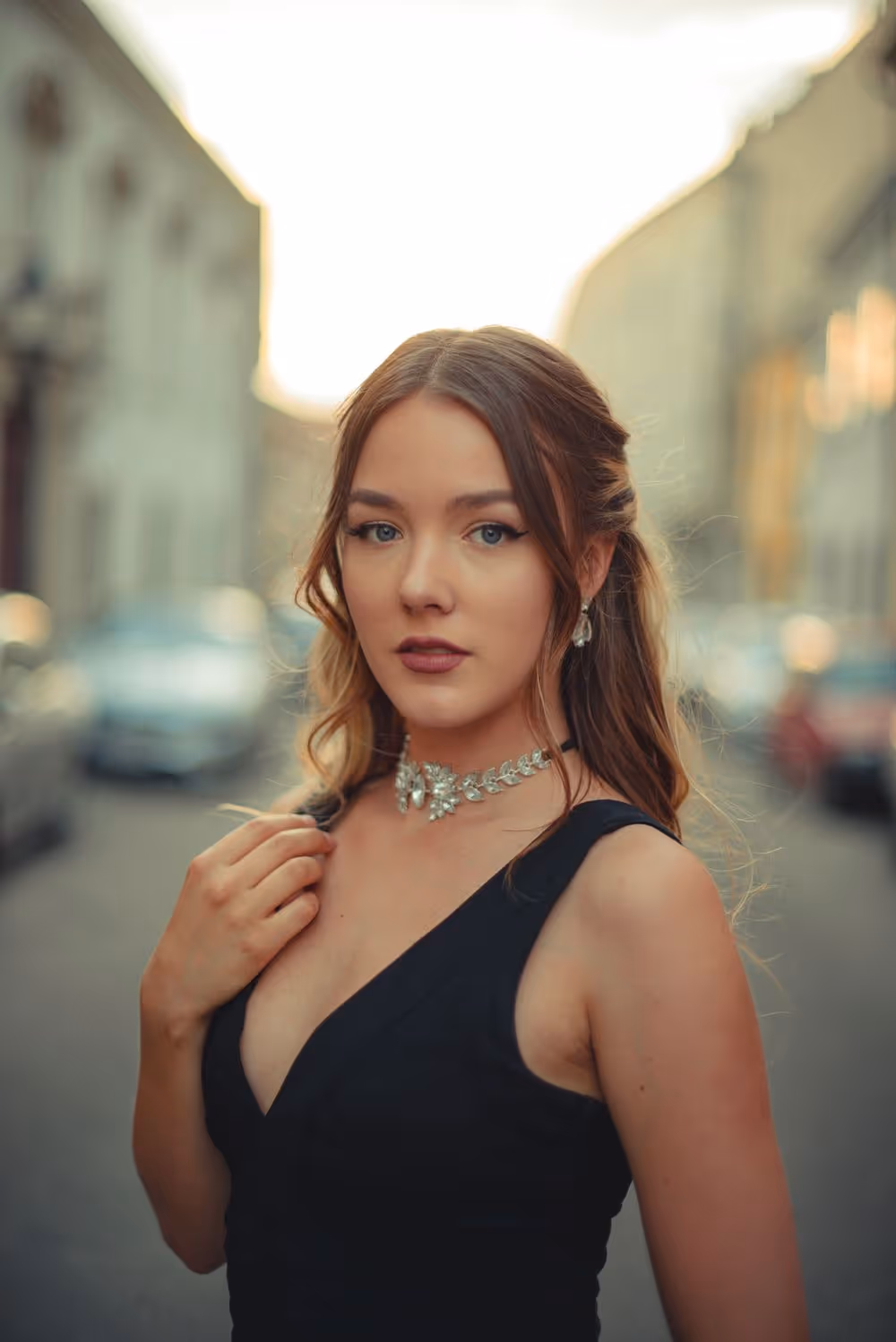 Young woman with blue eyes and long light brown hair wearing a black sleeveless dress and a silver necklace, standing on a blurred city street.