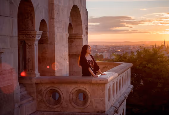 Woman standing on a stone balcony at sunset overlooking a cityscape.