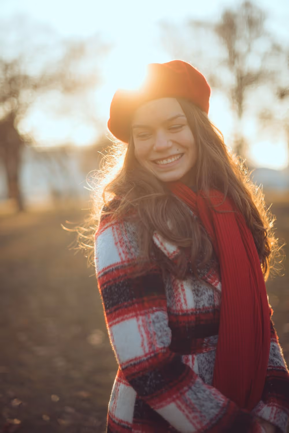 Smiling young woman wearing a red beret, red scarf, and plaid coat standing outdoors with sunlight behind her.