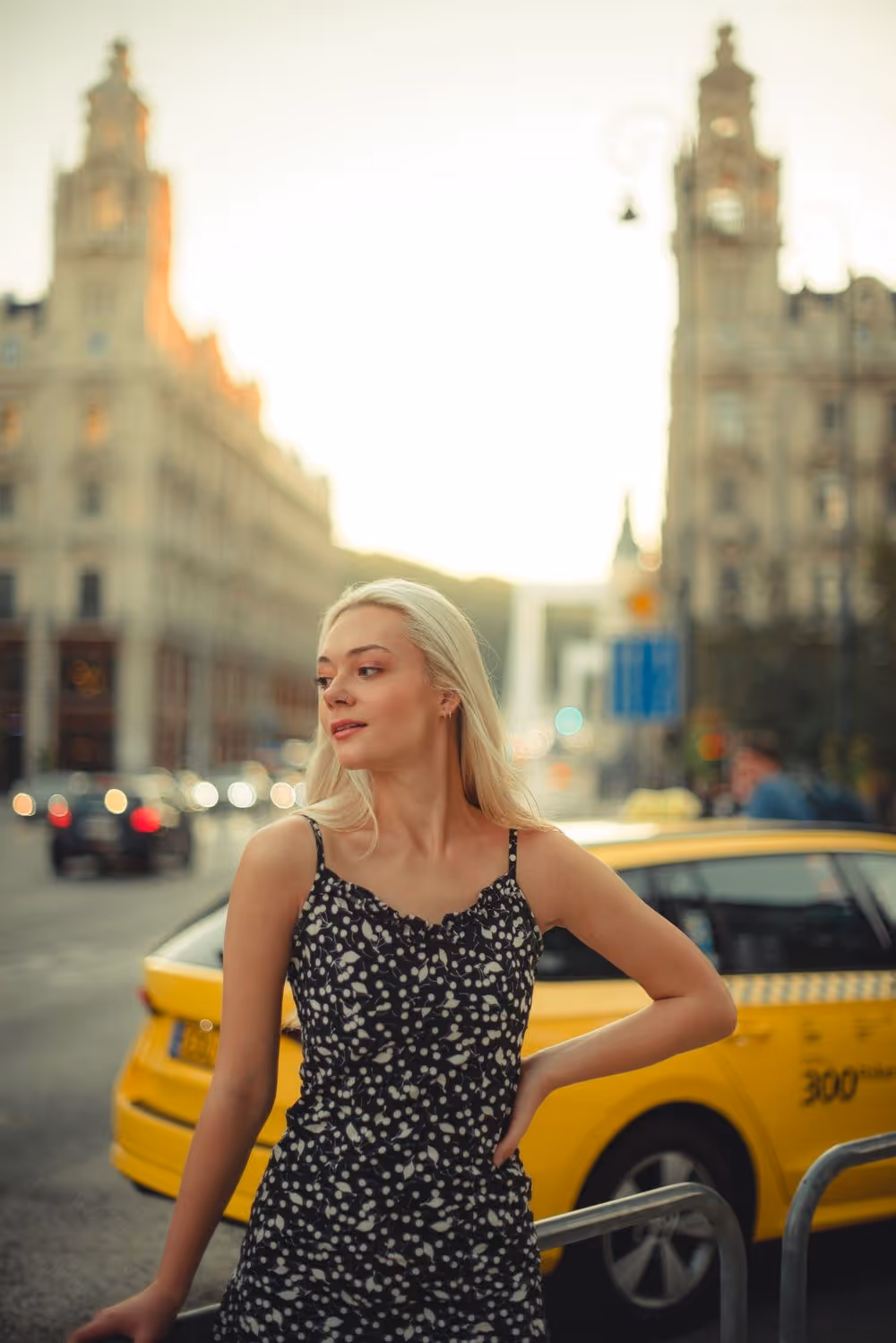 Young woman in a black and white patterned dress standing near a yellow taxi on a city street at sunset.