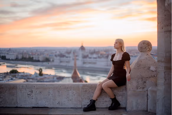 Young woman in a black dress and boots sitting on a stone ledge overlooking a city at sunset.
